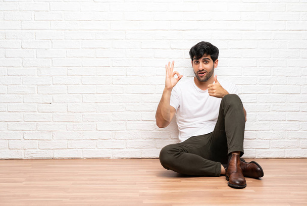 Молодий чоловік сидить на підлозі, показуючи знак ок і великий палець вгору жест
 - Фото, зображення