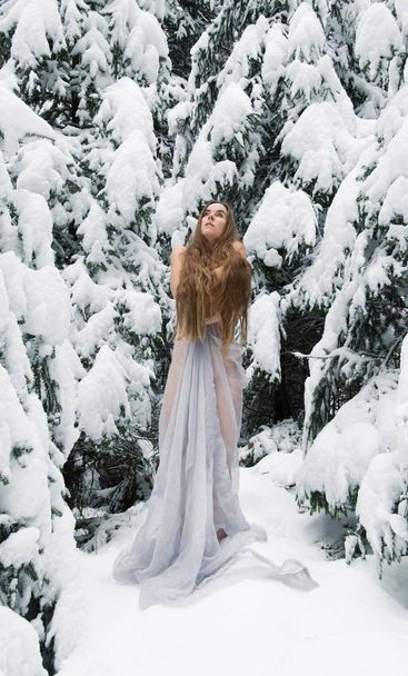 Jonge vrouw met lang haar, met lange witte jurk in de sneeuw. IJs bevriest in de winter, kijkt omhoog en warmt op met haar armen - Foto, afbeelding