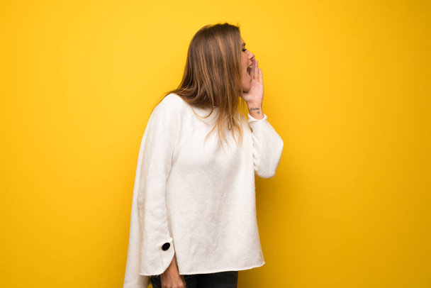 Mulher loira sobre a parede amarela gritando com a boca aberta para o lateral
 - Foto, Imagem