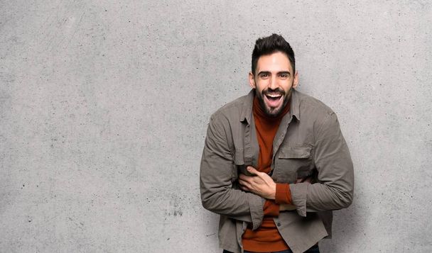 Handsome man with beard smiling a lot while putting hands on chest over textured wall - Photo, Image