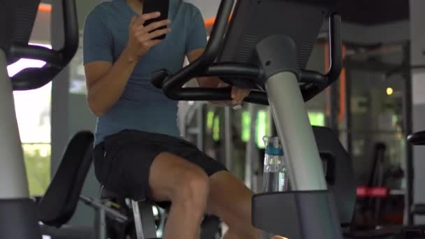 A young man at the gym on an exercise bike holding the phone in front of his eyes in his hands. All the attention is in the phone. The concept of dependence on social networks. Mobile addiction - Кадры, видео