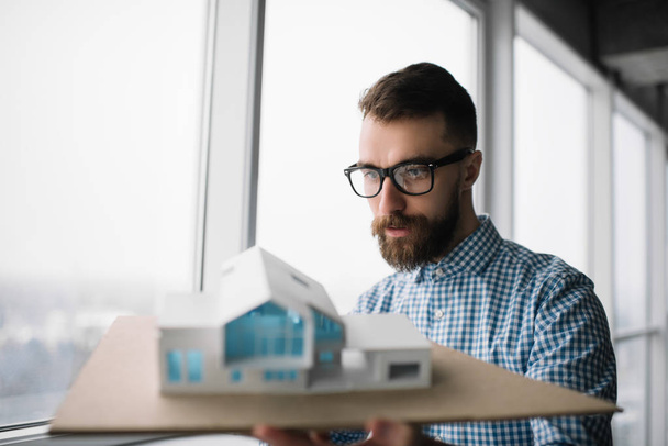 Young pensive architect holding a home model in loft modern office. Architectural project concept. University student planning engineering design, looking for solution, brainstorming at workplace - Foto, imagen