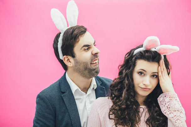 Young beautiful couple standing on a pink background. From the hare of the ears to the head. During this mans cheeks, having made a funny face looking into the camera. Easter concept. - Fotó, kép