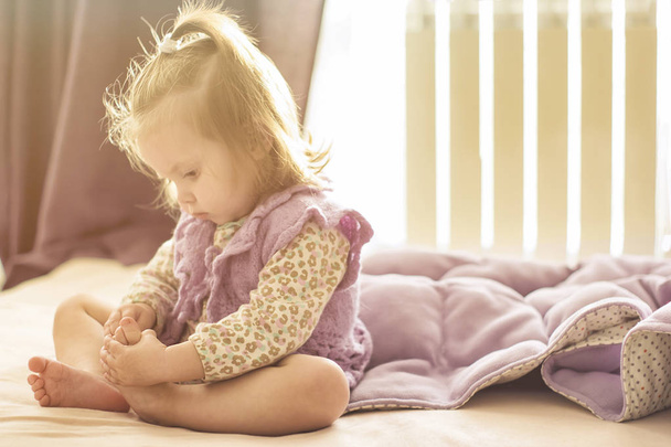Bir yatak Toddler kız sabah güneşte onu küçük ayaklar bakıyor üzerinde oturan kirişler - Fotoğraf, Görsel
