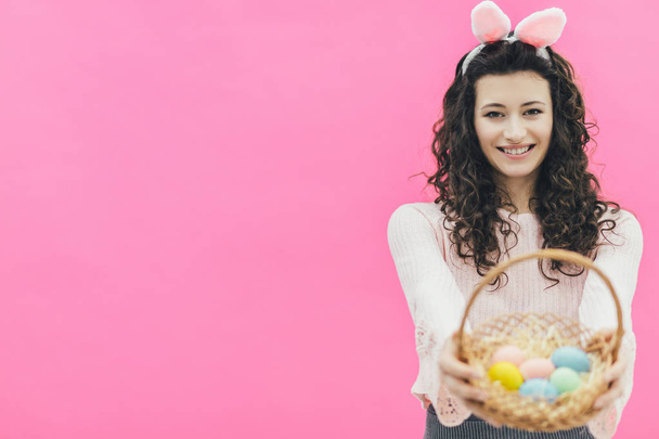 Young pretty girl standing on a pink background. There are bunny ears on the head. At the same time, he smiles and holds a basket of Easter colored eggs in his hands. Look into the camera. - Foto, Imagen