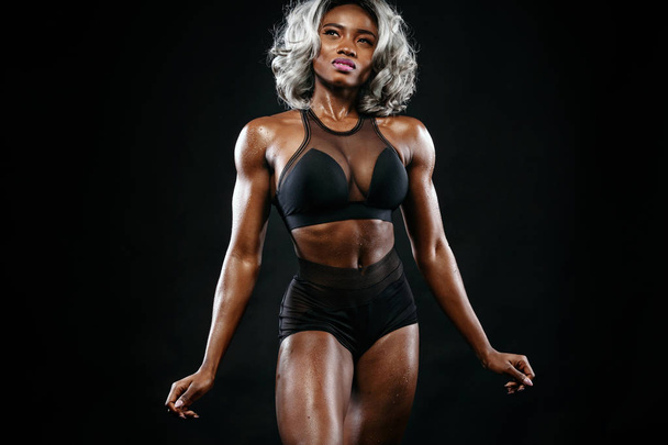 Sporty beautiful afro-american bodybuilder model, woman in sportwear makes fitness exercising at black background to stay fit - Fotografie, Obrázek