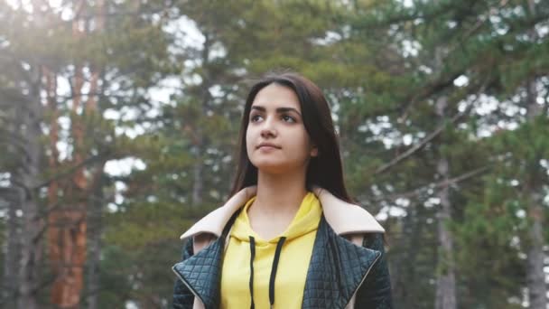 Romantic girl standing and looking around in pine wood in spring in slo-mo                                        Exciting view of attractive brunette girl with long loose hair in black and yellow jacket standing and looking around in wood in slo-mo - Filmagem, Vídeo