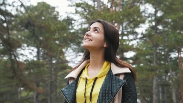 Smiling girl standing and looking around in fir forest in spring in slo-mo                                        Optimistic view of pretty brunette girl with long loose hair in  a black and yellow jacket hiking and looking in fir wood in slo-mo - Séquence, vidéo