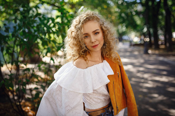 A beautiful young woman with blond curly hair walks through the sunny city - Photo, Image