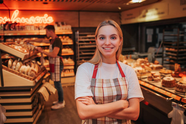 Happy young woman stand in grocery store. She pose nad look on camera. Happy worker smiling. Young man work behind. He put bread on shelf. - Foto, afbeelding