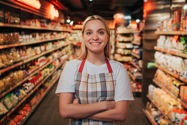 Busy young woman stand in line between shelfs with pasta. She pose on camera and smile. Happy positive model. - 写真・画像