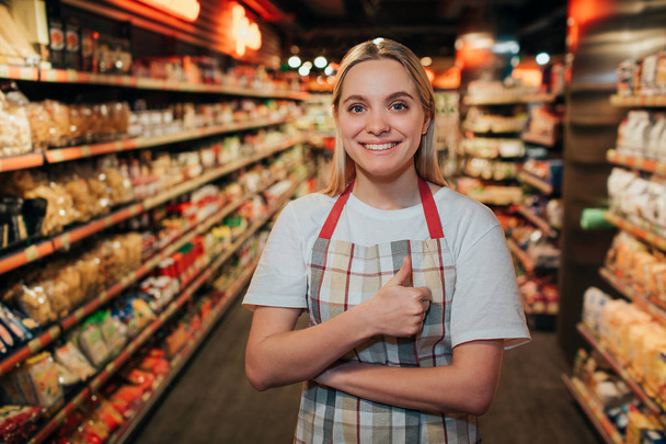 Happy positive young woman stand in line in grocery store. She look on camera and smile. Model keep big thumb up. - Foto, afbeelding