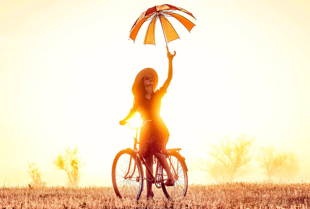Mädchen mit Regenschirm auf dem Fahrrad in der Natur bei Sonnenaufgang - Foto, Bild