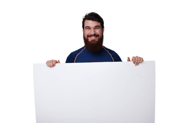 Front view. Portrait of smiling bearded guy holding blank white banner in his hands. Mock up, copy space for your text. - 写真・画像