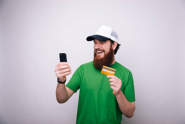 Görüntüsünü mutlu neşeli adam cep telefonuyla kredi kartı tutan liight gri duvar arka plan üzerinde izole ayakta sakallı. - Fotoğraf, Görsel