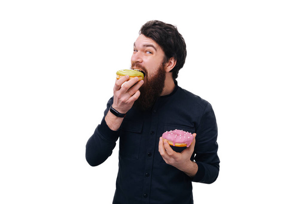 Hungry bearded man bites colorful  doughnut, opens mouth widely, isolated over white background, enjoys sweet taste - Photo, Image