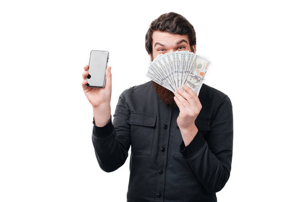 Photo of happy man in casual shirt holding fan of money in dollar banknotes and covering his face while showing blank screen mobile phone isolated over white wall  - Фото, изображение