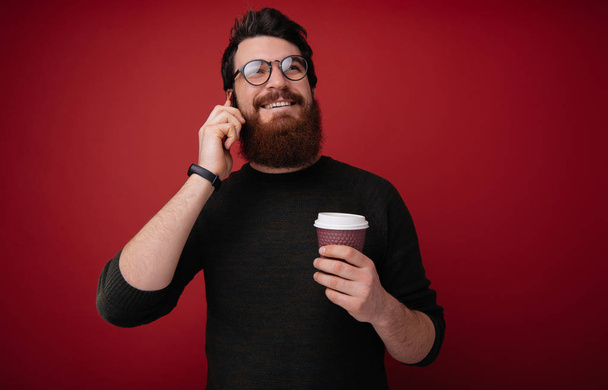 Красивый бородатый хипстер в очках, держит чашку кофе, разговаривая по смартфону
 - Фото, изображение