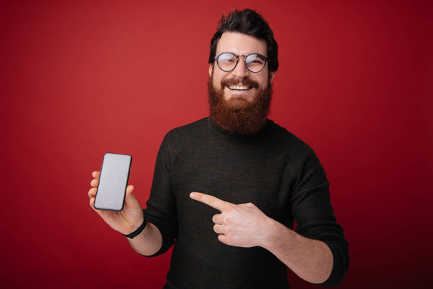 Εικόνα του νεαρή μελαχρινή γενειοφόρος άνδρας που δείχνει δάχτυλο στο smartphone και χαμογελά πάνω από κόκκινο φόντο απομονωμένες  - Φωτογραφία, εικόνα