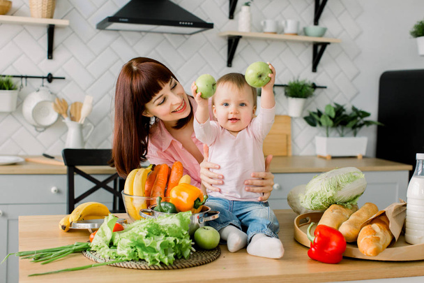 Happy laughing toddler girl and her beautiful young mother making fresh salad for breakfast together in a sunny white kitchen with a window - Фото, изображение