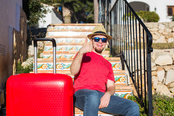 Tatil, seyahat, millet kavramı - bavul ile merdivenlerde oturan ve başparmak gösterilen genç adam - Fotoğraf, Görsel