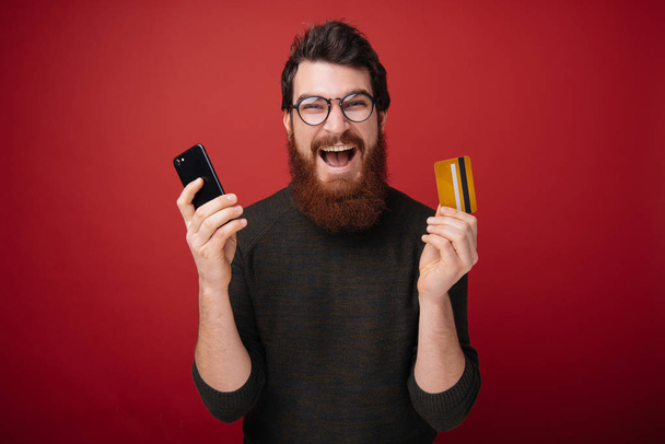 Photo de l'homme barbu excité émotionnel debout isolé sur fond de mur rouge tenant carte de crédit à l'aide d'un téléphone mobile. Caméra de regard
. - Photo, image