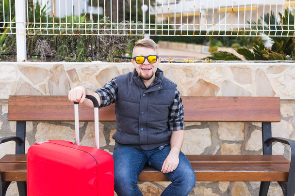 Reise, Tourismus und Menschen-Konzept - glücklicher Mann sitzt mit Koffer auf Bank und ist bereit für die Reise - Foto, Bild