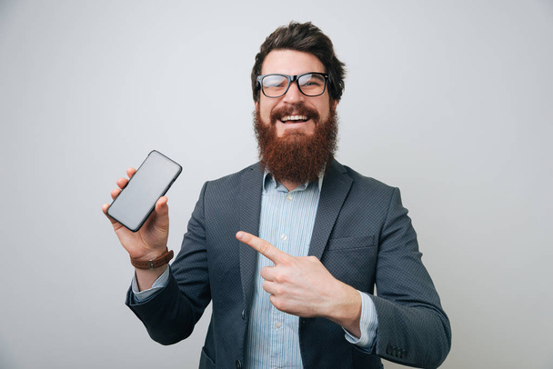 Retrato de hombre barbudo en ropa formal apuntando con el dedo en el teléfono inteligente y sonriendo en la cámara aislada sobre fondo gris
 - Foto, Imagen