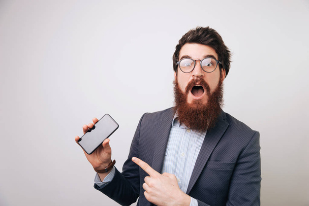 Bearded businessman with shocked face expression,  pointing at mobile over gray background - Foto, afbeelding