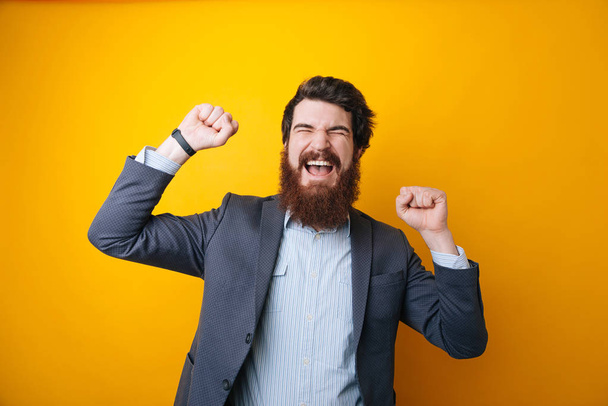 Foto di giovane uomo d'affari barbuto felice su sfondo giallo in piedi mentre fare gesto vincitore
 - Foto, immagini