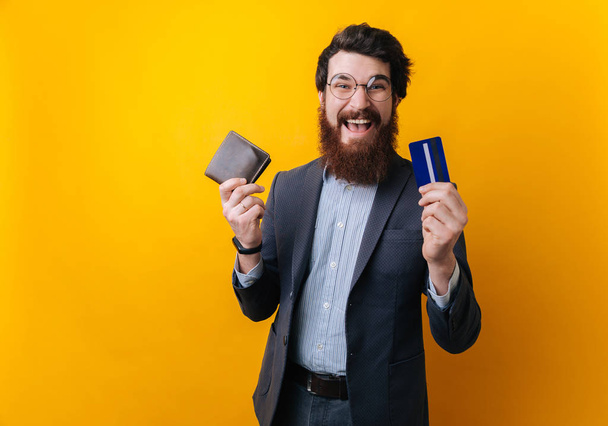 Image of excited bearded businessman holding old wallet and new credit card over yellow background  - Photo, Image