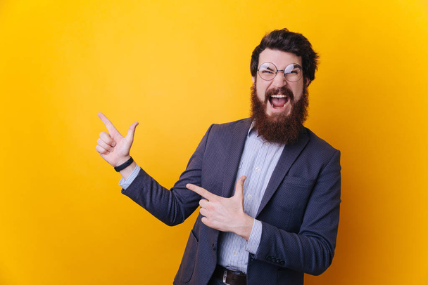 Onnellinen innoissaan parrakas mies silmälasit osoittaa pois kopioi tilaa kaksi sormea eristetty keltaisella taustalla
  - Valokuva, kuva