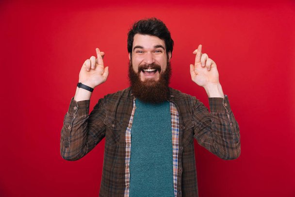 Молодой бородатый мужчина на изолированном фоне улыбается скрещивая пальцы с надеждой. Концепция удачи
. - Фото, изображение