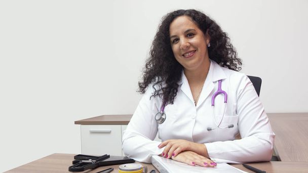 Doctora latina sentada sonriendo en su oficina con estetoscopio en el cuello sobre fondo blanco
 - Foto, Imagen