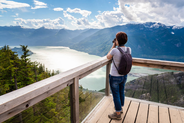 Mujer excursionista tomando fotos con una cámara DLRS desde una cubierta de visualización en la cima de una montaña con vistas a un fiordo. Impresionante paisaje en segundo plano. Squamish, BC, Canadá
. - Foto, Imagen