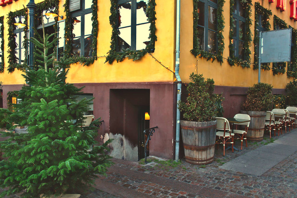 Canto de um edifício europeu amarelo com janelas decoradas com ramos de abeto para o ano novo e férias de Natal com um pinheiro verde na rua com uma ponte de paralelepípedos, mesas e cadeiras de um café de rua
 - Foto, Imagem