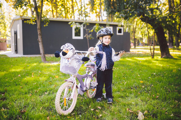 Funny dítě Kavkazský holka blondýnka v cyklistická přilba v blízkosti fialovým kolo s košíkem v mimo park na zelený trávník tráva vozíku doma. Přehraje používá rukou telefon s zubatými úsměv - Fotografie, Obrázek