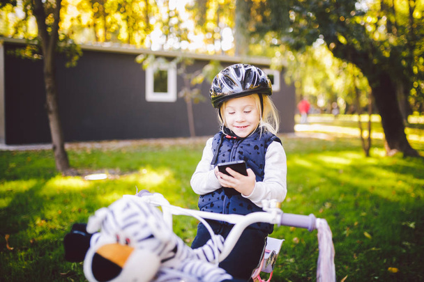 Смішні дитини кавказьких блондинка дівчина в велосипедний шолом поблизу purple велосипед з кошиком в за межами парку на зеленій галявині кошик травою на дому. П'єси використовує рукою телефон з зубастий посмішка - Фото, зображення