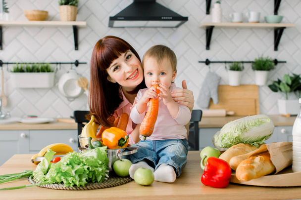 glücklich lachendes Kleinkind, das die Karotte isst und ihre schöne junge Mutter, die bereit ist, in einer sonnigen, hellen Küche frischen Salat für das gemeinsame Frühstück zuzubereiten - Foto, Bild