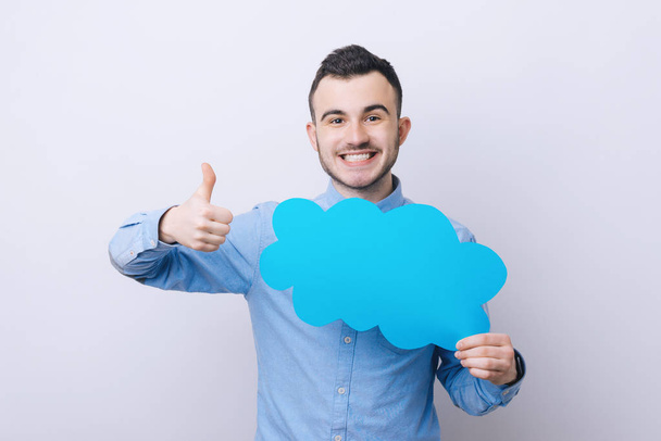 Handsome young guy is holding a speech cloud, gesturing  thumb up and smiling, on gray background - Photo, image