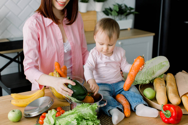 Cute little baby girl sitting on the table and holding carrot and green sweet pepper, helping her mother to prepare salad. Motherhood, food, healthy eating, cooking and family concept - Фото, изображение