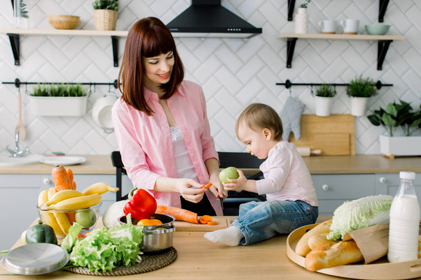 Smiling young mother chopping carrot while preparing fresh salad and little cute baby toddler holding green apple and sitting on the table in kitchen. Baby helps mother to cook at the home kitchen - Photo, Image
