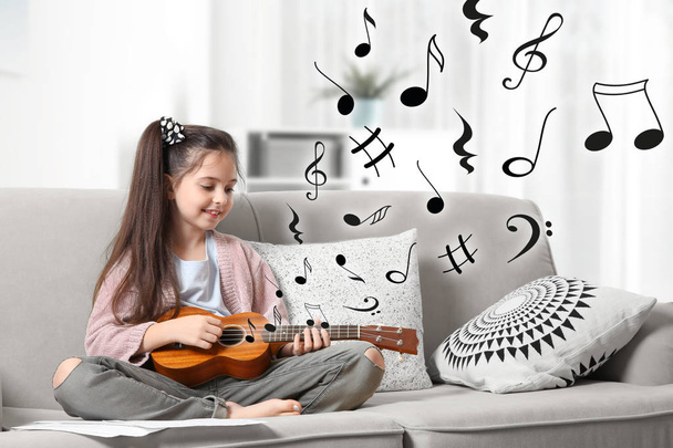 Cute little girl playing guitar on sofa and flying music notes in room - 写真・画像