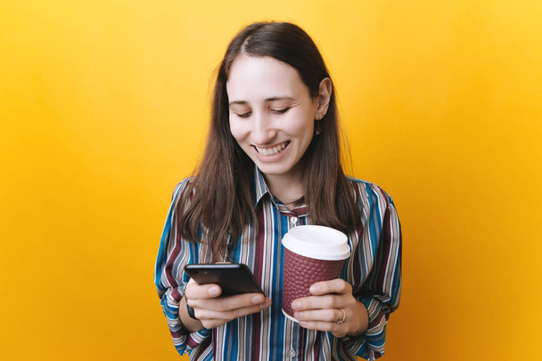 portrait of a smiling woman holding takeaway coffee cup and using mobile phone isolated over yellow background - Foto, Imagen