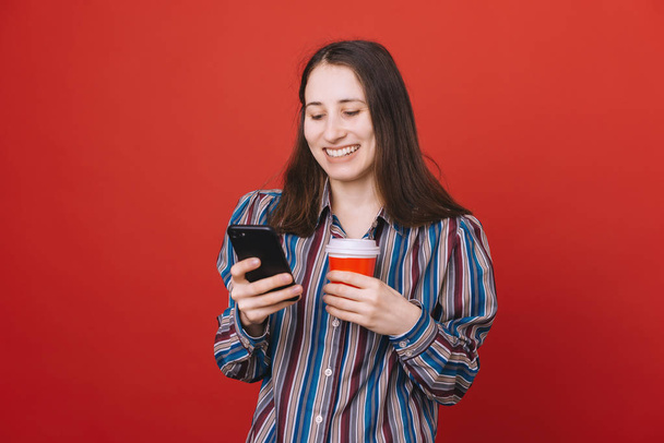 Retrato de uma jovem mulher casual isolada sobre fundo vermelho, segurando café takeaway usando telefone celular
 - Foto, Imagem
