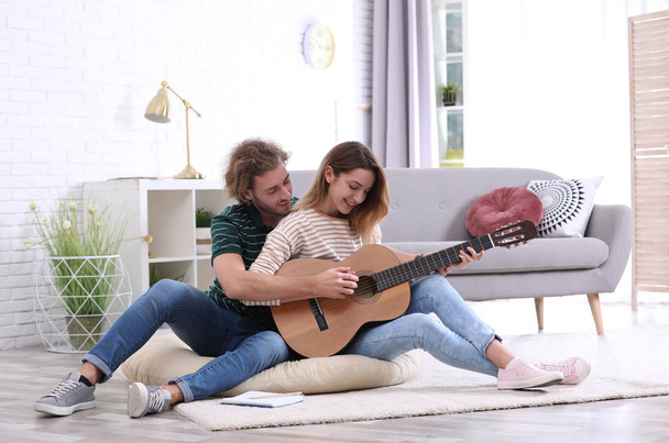 Young man teaching his girlfriend to play acoustic guitar in living room - Foto, Bild