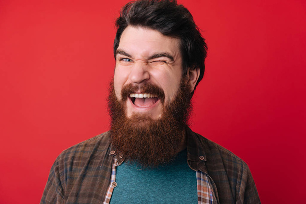 A handsome young bearded man  doing flirting gesture with eye over red background  - Fotoğraf, Görsel