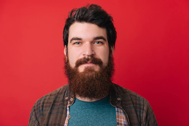 Tiro de joven guapo macho con barba, tiene expresión seria como escucha interlocutor, posa en estudio contra fondo rojo
 - Foto, imagen