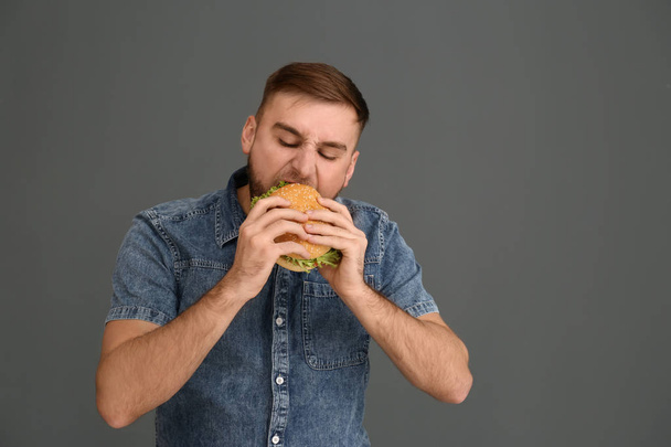Young man eating tasty burger on grey background. Space for text - Fotografie, Obrázek