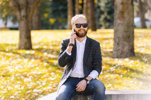 Hipster liikemies parta aurinkolasit hymyillen ja puhuva puhelin ulkona, istuu kaide
. - Valokuva, kuva